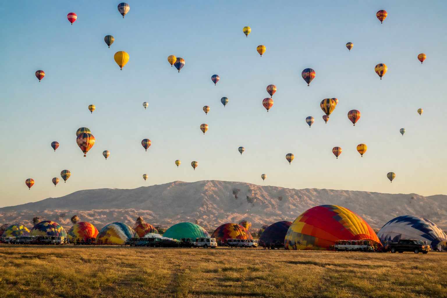 Dozens of hot air balloons in every colour rising from a wide field at sunrise, with more still inflating on the ground and mountains catching the first light behind