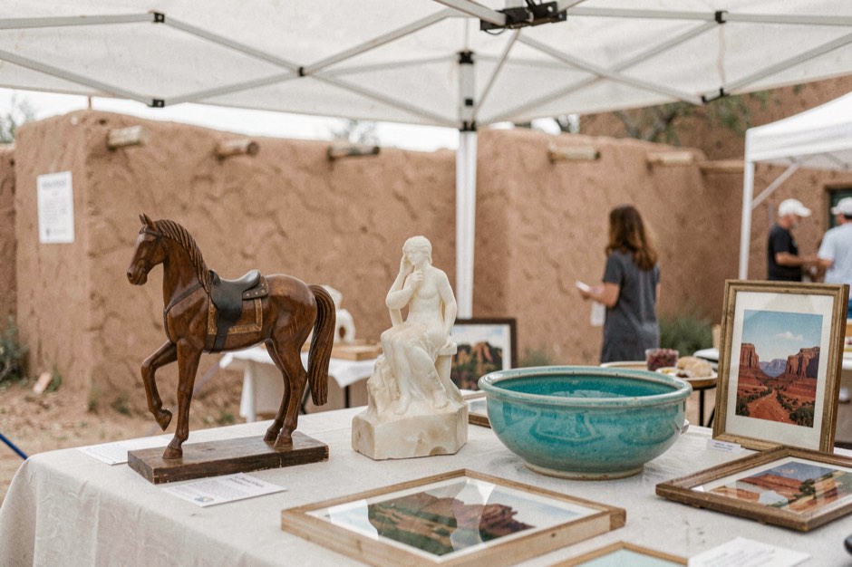 Close view of an artist's display table under a white canopy at an outdoor summer market, a hand-carved wooden horse, a polished alabaster stone sculpture, a turquoise-glazed ceramic bowl and small framed paintings of red mesas, adobe wall behind, soft daylight
