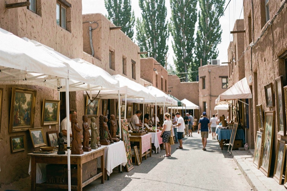 A narrow downtown street lined with white canopy booths on a bright summer morning, framed paintings and carved wooden sculptures on display tables, low adobe buildings and tall cottonwood trees behind, a few people browsing from booth to booth