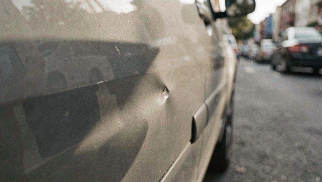 Close view of a shallow dent in the door of a parked car, soft light and reflections running across the dusty paint, with a quiet row of parked cars blurred further down the street.