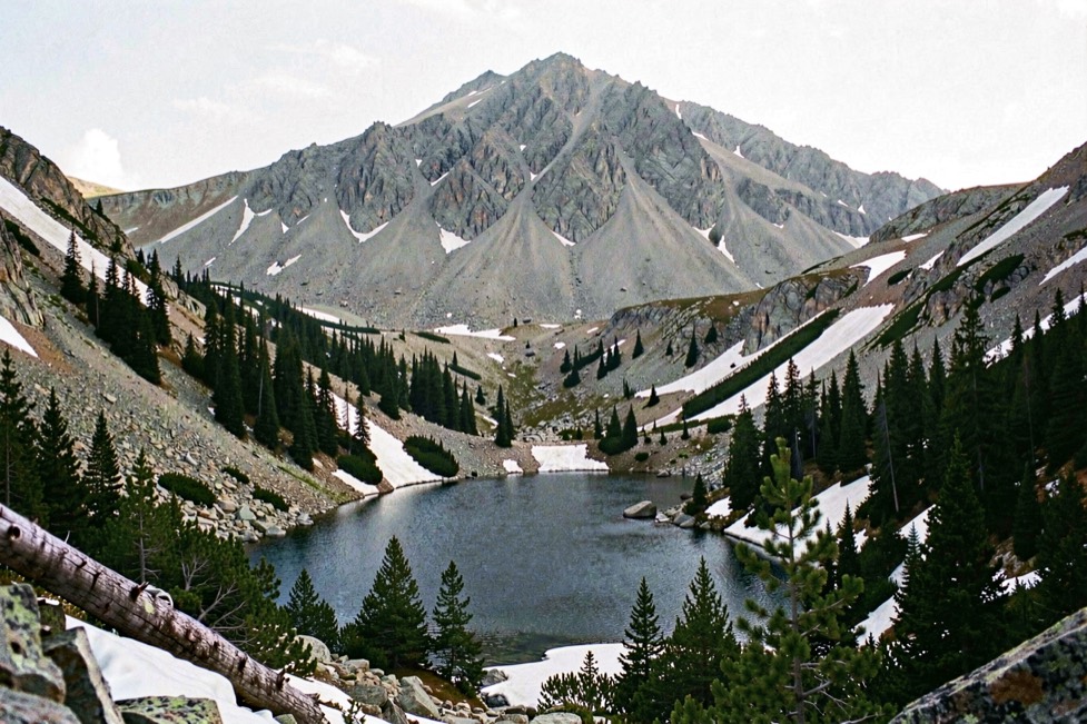 A small alpine lake in a rocky bowl below a bare grey summit ridge, dark spruce and fir along the shore and patches of old snow on the slopes, in clear morning light