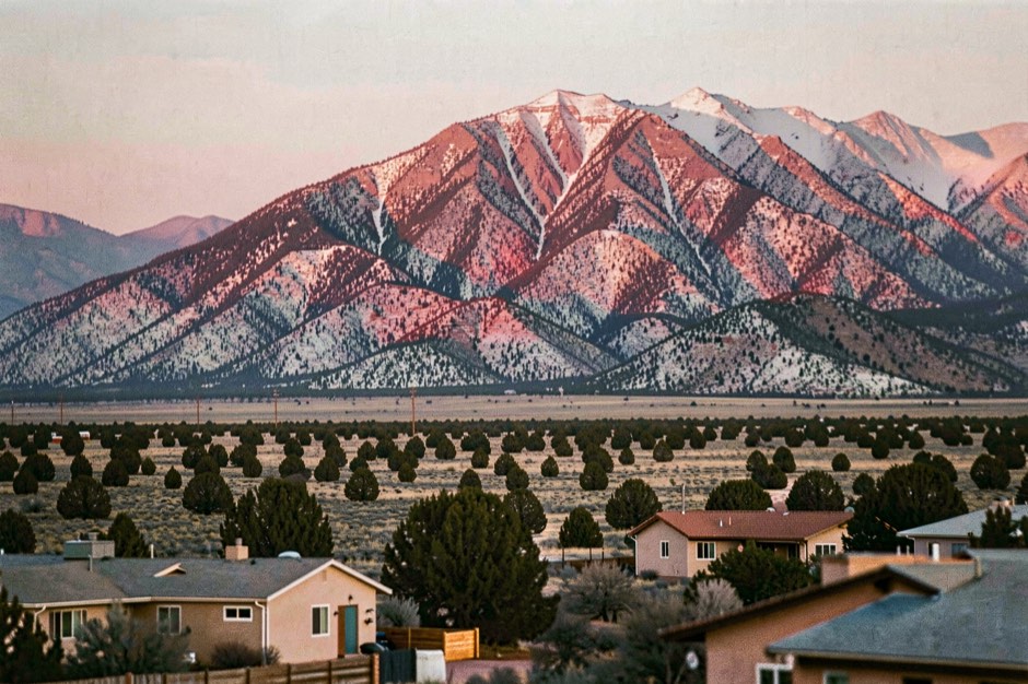 A long snow-dusted mountain ridge turning deep rose in the last evening light, above a high desert plain of scattered pi&ntilde;on and juniper with low earth-colored houses in the foreground